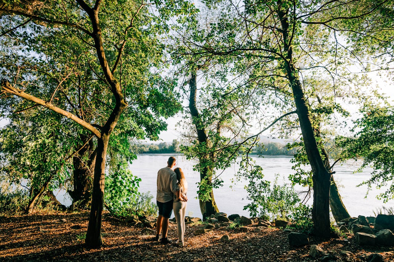 Couple embracing by the river surrounded by lush trees during a sunny day.